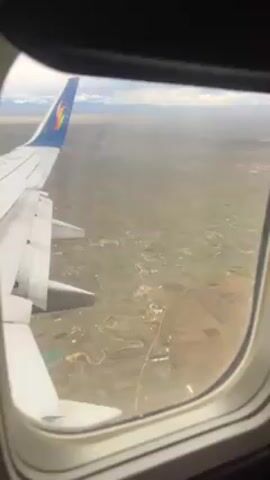 The video shows an aerial view from an airplane window. The airplane wing is visible in the foreground, with part of the plane's tail fin. Below, the landscape features fields of green and brown. Roads and a small winding river are present. Some scattered buildings are visible. In the distance, there are mountains and a cloudy sky.