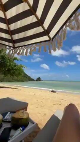 The video shows a tropical beach scene. It starts with a view from under a striped umbrella with white fringe, overlooking a tan sandy beach and calm, turquoise water with a rocky outcrop in the distance. Lush green vegetation borders the beach. A chaise lounge with a coconut drink and phone on the side table is in the foreground. The camera moves to show a woman in a floral bikini and sunglasses relaxing on a lounge chair covered with a teal towel.  Palm trees and other beach umbrellas are visible in the background. The camera is held by someone, possibly a friend, who is partly visible in the frame.