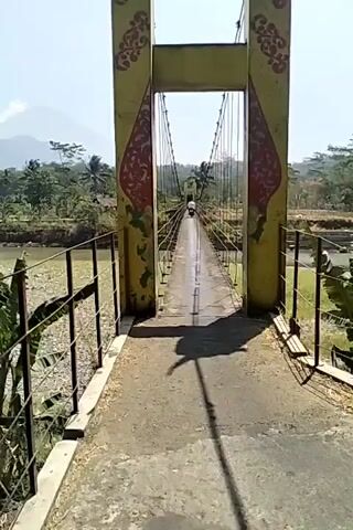 The video shows a view from a suspension bridge, looking down its length. The bridge is supported by two tall, yellow pillars decorated with red and yellow patterns. The bridge deck is made of concrete and appears wet in places. A single person is visible riding a motorcycle in the distance, in the center of the bridge. The surrounding environment includes a dry riverbed with rocks and vegetation, trees, and a mountain or large hill in the background. The sky is clear and blue, indicating a sunny day. The camera appears to be handheld and moving slightly, suggesting the person filming is walking or riding across the bridge.
