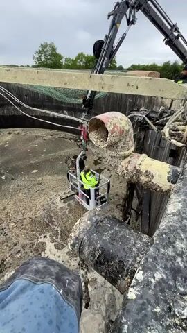 The video shows two individuals in a man lift being lowered into a circular basin of a milky, foamy liquid, presumably wastewater. The lift is suspended by a crane. The inside walls of the basin are concrete. There is a safety net stretched along the upper part of the basin. A large, old pipe runs along the edge of the basin where the camera is mounted. The surrounding environment is green and cloudy, indicating an outdoor industrial setting. One of the individuals is wearing a bright yellow reflective jacket.