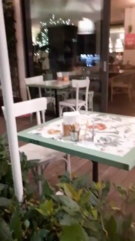 The video shows an outdoor seating area of a restaurant at dusk or night. Tables with white chairs are set up, some with green-trimmed tabletops. Plants in planters line the sidewalk and the seating area. People are seated at tables inside the restaurant, visible through large windows. Some people are walking along the sidewalk outside. The lighting is dim, with string lights visible in the background.