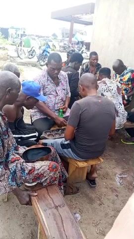 A group of men are gathered outdoors, seated on wooden benches and stools, playing a board game. The game board, with its characteristic rows of depressions, is visible in the center of the group, and a green bottle sits next to it. The men are dressed in casual clothing, with some wearing traditional attire. The environment appears to be a dirt or sandy area, with a building in the background and some motorcycles parked in the distance. The scene suggests a casual social gathering, possibly a game of strategy or chance.