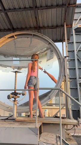 A young person with blonde hair tied in a ponytail and wearing a pink tank top and denim shorts stands on a metal platform. They are reaching out their right hand towards the reflection of another person's arm and hand in a large, circular, mirrored object. The mirrored object is part of a structure that appears to be industrial or scientific. The background shows a sky and the tops of buildings, suggesting an outdoor setting. The lighting indicates it may be late afternoon or early evening. The scene is framed from a slightly low angle, looking up at the person and the mirrored structure.