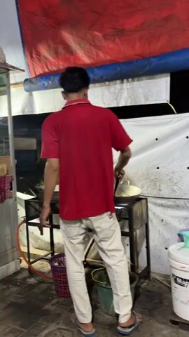 A person wearing a red shirt and light-colored pants is standing and cooking in front of a stove. They are holding a utensil and stirring something in a pan. The background shows a tarp, a white wall, and various containers. The scene appears to be an outdoor or semi-outdoor setting, possibly a food stall or market.
