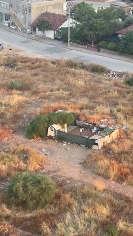 A small, makeshift dwelling made of corrugated metal and tarps is situated in a dry, grassy field. The dwelling has a fenced-in area in front of it. A person, possibly a child, is visible inside the fenced area. In the background, a street lined with houses and trees can be seen. The overall impression is one of a humble, possibly improvised, living situation in an undeveloped or rural area.