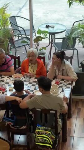 A group of six people are seated around a table, which is covered with a black and white checkered tablecloth. The table is laden with bowls of food, plates, glasses of red liquid, and possibly some cutlery. On the left side of the table, an older man in a pink and blue striped shirt is leaning on his left hand, looking down. Next to him, an older woman in an orange jacket over a yellow top is looking forward and appears to be speaking. To her right, a woman with dark hair is resting her chin on her hand, looking down. In the foreground, facing away from the camera, a man in a light green shirt is seated at the table. To his left, a young boy in a blue shirt is facing the man, with his back to the camera. To the left of the boy, a woman in a white top is leaning forward, facing the young boy. The environment appears to be an indoor seating area with large windows in the background, showing outdoor seating with tables and chairs, and lush green plants. Some of the chairs have backpacks on them. The lighting suggests it is daytime.