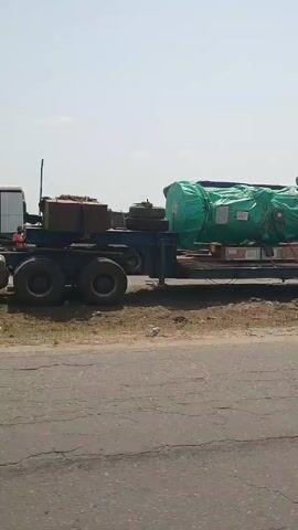 The video shows several tractor-trailers parked on the side of a road. In the first shot, a trailer is carrying a long object, wrapped in green tarpaulin and secured with chains. The camera then pans to the right, showing a red semi-truck with the number 67 on its door, parked in front of a blue shipping container on another trailer. The video continues, panning to the right to show another white semi-truck, then another flatbed trailer carrying a large object covered in blue tarpaulin. In the background, there are large metal structures and a crane. The environment appears to be an industrial or port area, with a cracked asphalt road in the foreground and a clear sky.