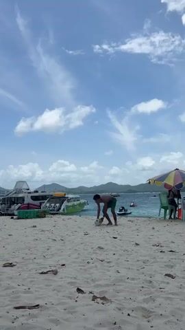 A young man in green shorts is playing soccer on a sandy beach. He is barefoot and has dark hair. In the background, there are several boats docked on the water, with mountains and a cloudy blue sky behind them. The man kicks the ball, then runs after it. The camera follows him as he moves along the beach.