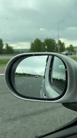 The shot is taken from inside a moving car, looking at the passenger side mirror. The mirror reflects a highway with other cars, under an overcast sky. Trees line the side of the road. The mirror itself has water droplets on it.