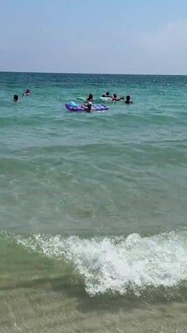 A group of people are swimming and relaxing in clear turquoise water near a beach on a sunny day. Some individuals are floating on inflatable rafts, while others are swimming in the ocean. The water is shallow enough near the shore to see the sandy bottom. The sky is clear and blue, with no clouds visible. The scene appears peaceful and recreational.