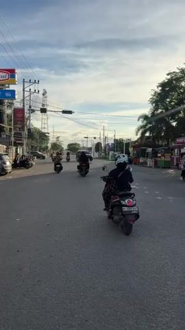 A vertical, medium shot captures a street scene with several motorcycles and a car on a paved road. The sun is low in the sky, casting long shadows. On the left side of the road, there are buildings, including a convenience store with a red and blue sign that reads "Indomaret." Several motorcycles are parked in front of the buildings. In the center of the road, a person is riding a motorcycle, and a black car is driving in the same direction. On the right side of the road, there are food stalls and more trees. The sky is blue with scattered clouds. The overall atmosphere is that of a typical day in a residential or commercial area.