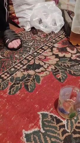 The video shows a person's foot wearing a sandal on a carpeted floor. Various items are scattered around, including plastic bags, shoes, and a partially filled bottle of amber liquid. A small glass with a similar amber liquid is also visible on the carpet. The background features a wall covered with patterned wallpaper. The scene appears to be indoors, likely a living space.