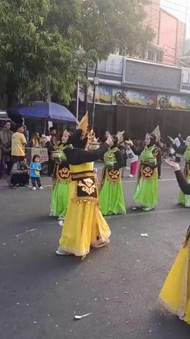 A group of dancers in traditional yellow and green costumes with ornate headpieces are performing in a street parade. The dancers are holding small, decorated bowls. Several onlookers, including children and adults, are watching the parade from the side of the street. The background shows buildings and trees. The overall atmosphere appears festive.