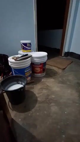 The video shows a low-angle view of several buckets and paint cans on a concrete floor next to a doorway. The room appears to be in the process of being painted or renovated. In the foreground, a black bucket contains a white liquid, presumably paint or primer. To the right of the black bucket are two large white paint buckets. One is labeled "BERGER Robbialac CLASSIC ACRYLIC PREMIUM EMULSION" and features a red lamp graphic. The other, partially obscured, has blue and yellow labeling with the brand name "BERGER" and the text "WATER". Behind these, a smaller white paint can with blue and yellow branding is stacked on top of another white lid. A trowel or putty knife rests on the lid of the "BERGER Robbialac" bucket. The floor is dusty and shows signs of wear. The background reveals a dark, open doorway leading to another room. A small brown doormat is visible near the doorway. The lighting is dim, suggesting either low ambient light or that the video was taken at night.
