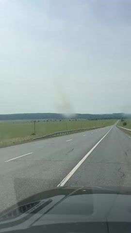 A first-person perspective video of a car driving on a paved road through a rural landscape. The road is asphalt with a white line down the center and a white guardrail on the right side. The surrounding environment consists of green fields and trees, with a hazy sky. There are no other vehicles or people visible.
