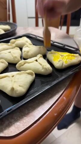A close-up shot shows a person's hands preparing uncooked pastries on a baking sheet. The pastries are triangular with a folded-over top, revealing a dark filling. A hand is seen holding a small brush dipped in a bright yellow liquid, likely egg wash, and is in the process of brushing it onto one of the pastries. Another hand holds a small, clear plastic container filled with the same yellow liquid. The baking sheet is black and sits on a table with a wooden edge. In the background, chairs and cabinetry are partially visible, suggesting an indoor kitchen setting. The lighting is warm, highlighting the food preparation activity.