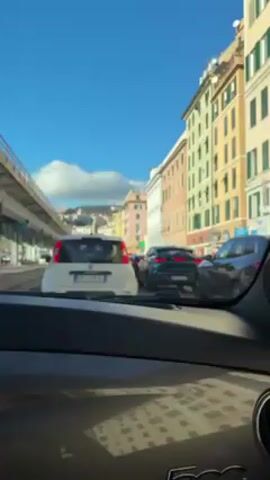 The video shows a view from inside a car, focusing on traffic congestion on a city street. Several cars are visible, including a white Fiat Punto, a dark green Maserati Grecale, and a dark-colored SUV. The buildings lining the street are painted in various pastel colors and the sky is clear and blue.  An elevated highway overpass can be seen to the left of the road. The dashboard of the car from which the video is recorded is visible in the foreground.