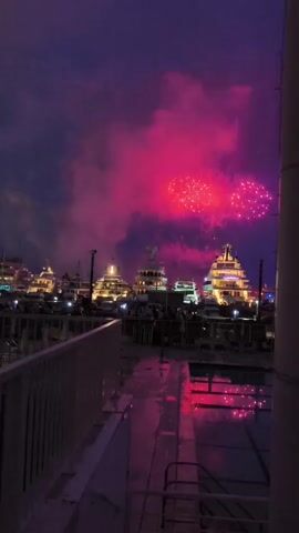 The video captures a nighttime scene in a harbor. Several large yachts are docked, illuminated with warm lights. The sky is filled with vibrant pink and red fireworks, creating a dramatic effect. Smoke from the fireworks billows across the sky. In the foreground, a railing and part of a deck or walkway are visible, with reflections of the fireworks and yacht lights shimmering on a wet surface, possibly rain-slicked pavement or water. The overall atmosphere is festive and celebratory. No individuals are clearly distinguishable.