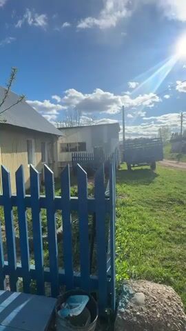 The video shows a view from behind a blue picket fence in a rural setting on a sunny day with scattered clouds. To the left of the fence, there is a partially visible, light-colored house with a metal roof. A dark blue trailer sits in the grassy yard next to a dirt road. The road leads past various buildings, including a brick structure with a red roof and a small, older house, under telephone poles and wires. A silver car drives down the dirt road in the distance. The sun is bright and creates a lens flare effect. A small bucket sits near the base of the fence. The camera slowly pans to the right, revealing more of the grassy area and the dirt road.