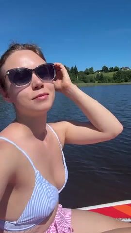 A young woman with sunglasses and a bikini top is sitting on a paddleboard in a lake. She is wearing a striped bikini top and pink and white striped shorts. She is holding her hand to her forehead, shielding her eyes from the sun. The water is calm and the sky is blue. The background shows trees and a distant house.
