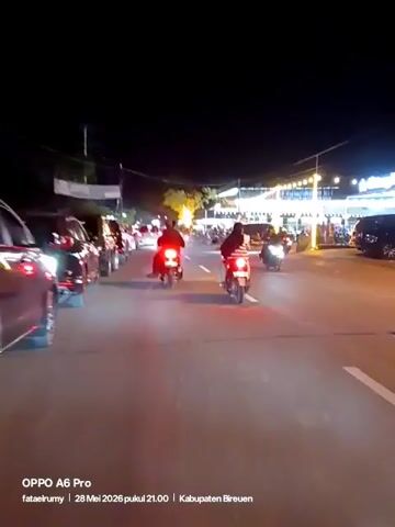 The video shows a night scene of a street in Kabupaten Bireuen, Indonesia, filmed on an OPPO A6 Pro on May 28, 2026, at 9 PM. Several cars and motorcycles are present on the road, with a line of parked vehicles on the side. There appear to be restaurants or businesses lit up in the background. People can be seen riding motorcycles and walking around. The road is marked with white lines.
