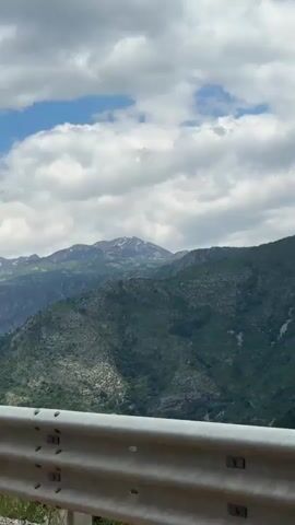 The video shows a mountain range under a cloudy sky. The mountain in the distance has a dusting of snow on its peak. The foreground hills are covered in green vegetation and exposed rock. A guardrail is visible at the bottom of the frame. The camera zooms in on the mountain.