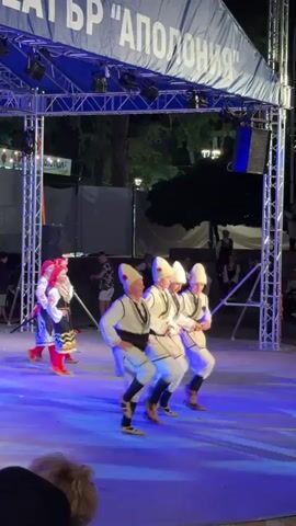 The video shows a performance on an outdoor stage at night. Several individuals in traditional folk costumes are dancing on a brightly lit blue stage. The costumes appear to be white with black and red accents. Four male dancers in the foreground are performing a synchronized dance, with one man in the center raising his arm. Behind them, three female dancers in similar, though distinct, traditional attire are also dancing. A large blue banner with white lettering, which reads "ТЕАТЪР 'АПОЛОНИЯ'", is visible above the stage, illuminated by spotlights. The background shows trees and lights, suggesting an urban or park setting at dusk or night. There are spectators visible in the lower foreground, partially out of focus.