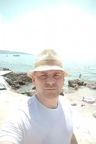 A man wearing a white t-shirt and a straw fedora hat is taking a selfie at a beach. He is standing on a rocky shoreline with the ocean in the background. There are several people in the water swimming and playing. The sky is bright and sunny. The man is looking directly at the camera.