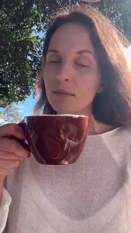A woman with long brown hair and a white knitted sweater is holding a brown mug of coffee. She brings the mug to her lips and takes a sip, then pulls it away and sticks out her tongue briefly. The background shows green trees and a blue sky.