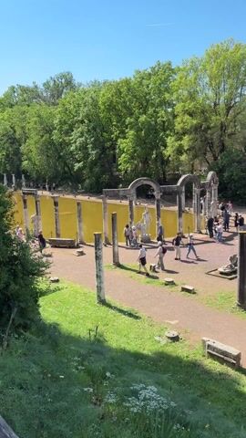 The video shows a scene in what appears to be an archeological site. There is a large pool of water, tinted yellowish-brown, with partial column structures and statues surrounding it. Numerous people are walking around the area, appearing to be tourists exploring the site. There are several columns scattered throughout the open space, and in the background is a brick building or structure with multiple windows. The day is sunny with a clear blue sky, and there are trees providing shade around the edges of the area. The ground consists of dirt pathways and patches of grass. The video seems to have been shot from an elevated position, offering a broad view of the historical site.