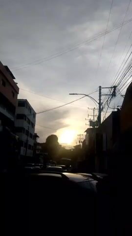 The video is a vertical shot of a street with buildings on both sides and cars parked or driving in the foreground and middle ground. The sun is setting, casting a warm glow and creating a silhouette effect on the buildings and vehicles. The sky is overcast with thin clouds, and there are power lines visible against the sky. The overall impression is a street scene at dusk.