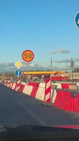 The video shows a view from inside a vehicle, looking forward onto a road. The road has been narrowed by temporary barriers, which are a mix of red and white striped delineators and white and red plastic water-filled barriers. There are several traffic signs visible. Closest to the camera, there is a diamond-shaped yellow sign with a white border, indicating a priority road. Below that is a circular blue sign with a white arrow pointing upwards, indicating a straight-ahead direction. To the right, a large circular red sign with a white border contains the number "50" in yellow, indicating a speed limit of 50. Further down the road, another similar set of signs is visible, including a speed limit of 50. Other vehicles are visible on the road ahead. In the background, there is a gas station with a yellow canopy and red pumps, as well as some trees and buildings. The sky is blue with some scattered clouds. The overall impression is that of a road with ongoing construction or traffic management.