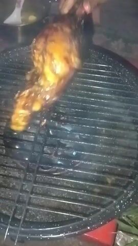 The video shows a person grilling chicken on a small, round grill. The chicken is glazed with a dark, glossy sauce. The person is using tongs to maneuver the chicken on the grill. A bowl containing liquid sits in the background. The grill is located indoors on top of what appears to be a red plastic bin.