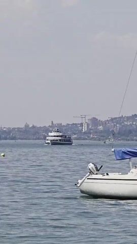 The video opens with a distant shot of a large passenger boat moving across a body of water, with a city skyline visible in the background. The camera then pans slightly to the right, revealing a white sailboat in the foreground with a blue cover over its boom. More buoys are visible in the water, and the city skyline continues to stretch across the horizon. As the video progresses, the camera continues to pan and zoom out, showing more sailboats and docked boats, some covered with tarps. The water appears somewhat choppy. The sky is a hazy blue, with some clouds. The video concludes with a wider view of the lake, showing numerous boats moored and anchored, with a clear blue sky overhead. The shore is visible on the left side of the frame, with some concrete and rock formations.