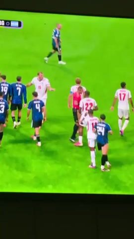A group of male soccer players in white and red uniforms are gathered on a vibrant green field. One player in a red referee's uniform is in the center of the group, facing a player in a white uniform with the number 9 on his back. The referee is holding his left arm up, seemingly gesturing or giving an instruction. Other players in white uniforms are positioned around the central figures, some looking towards the interaction, while one player in a blue uniform with the number 24 on his back walks away from the group. The background is blurred green, indicating a sports field.
