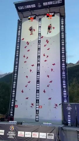 This is a video of a speed climbing competition. Two male athletes are climbing up a very tall, artificial climbing wall. They are side-by-side, each in their own lane. The wall is white with red climbing holds. There are digital timers at the top of the wall displaying their times. The environment appears to be outdoors, possibly at a mountain resort, as indicated by the trees and mountains in the background. The competition appears to be part of a "World Climbing" event, as suggested by banners at the bottom of the screen. The athletes are wearing athletic clothing and harnesses. The wall has markings on the side indicating height markers and possibly lane designations. The lighting suggests it is either early morning or late afternoon.