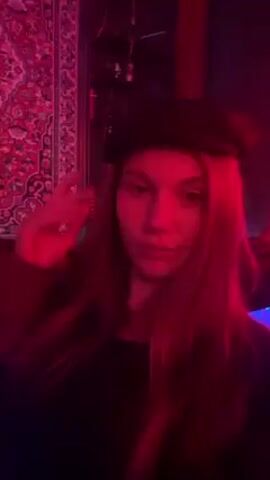 A young woman with long brown hair is shown in a dimly lit room bathed in red light. She is wearing a black top and a dark beret. The woman faces the camera, smiles, and tips her beret. Behind her is a patterned tapestry on the wall and some indistinct objects. The overall atmosphere is that of a dimly lit bar or club. Music is playing in the background.