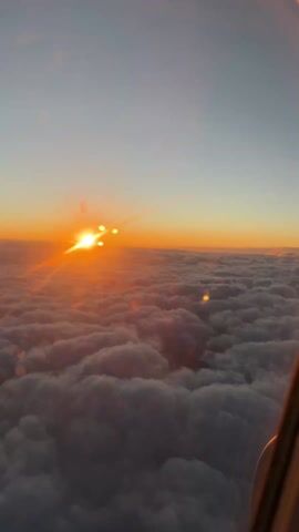 The video shows a sunset above a thick cloud layer, likely taken from an airplane. The sun is positioned on the horizon, appearing as a bright orange-yellow orb. Its light reflects and spreads across the cloud cover, creating a warm, vibrant glow. The sky transitions from a pale blue to a deeper orange near the horizon. Part of the plane's interior is visible in the bottom right corner.