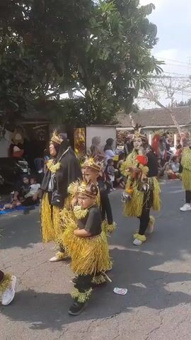 A group of people, including adults and children, are walking in a parade. They are dressed in elaborate costumes made of straw or raffia, with headdresses and ornate collars. The costumes are predominantly yellow and black. Some of the participants are carrying props, such as sticks with colorful decorations. Spectators are lining the street, watching the parade. The environment appears to be a residential area with houses and trees. The weather seems to be sunny.