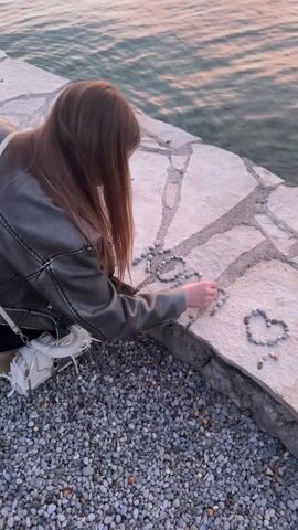 A young woman with long reddish-brown hair is kneeling on a stone ledge by the water. She is wearing a gray leather jacket, black pants, and white sneakers, with a white purse slung over her shoulder.  She is arranging small, gray pebbles on the stone surface to form the word "LOVE" and a heart shape. The water is visible behind her, and a large area of small pebbles surrounds her on the ground. The lighting suggests it is either sunrise or sunset.