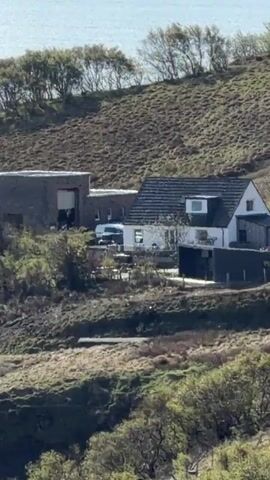 The video shows a house nestled into a hillside near a body of water. The video zooms out to show the whole body of water with hills surrounding it, taken on a sunny day with light clouds. A number three is on a marker in the grass with large rocks in the foreground.