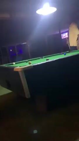 Here is a description of the video.
The video shows a person in a yellow shirt standing near a pool table, holding a cue stick. The pool table is green with several balls on it. A bright light hangs above the table. The floor around the table appears to be artificial turf. In the background, there is a building with a blue light inside. The camera is angled up at the pool table. Another person can be partially seen at the edge of the frame, also holding a cue stick.