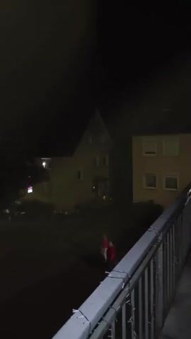 The video shows a dimly lit outdoor scene at night. There are buildings, parked cars, and trees visible. A person wearing a red and white outfit, possibly a costume, is walking across the area. The camera appears to be mounted on a balcony or elevated platform, looking down on the scene. There is a railing in the foreground. The lighting is minimal, with some streetlights and lights from the buildings illuminating the area. The atmosphere is quiet and still.