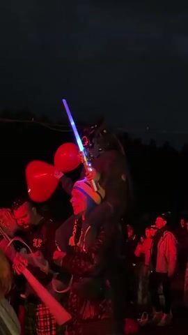 A video captures a man giving a child a shoulder ride at night. The man wears a blue and yellow beanie and a blue jacket. The child, wearing a dark jacket and a Predator-style mask with red glowing eyes, holds two red balloons in one hand and a blue light-up toy sword in the other. Another person, partially visible on the left, also holds a blue light-up sword. The background is dark with faint hints of people and trees. The child raises the sword and balloons in the air.
