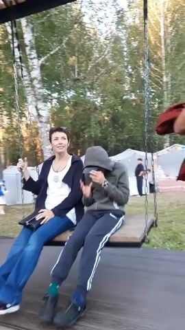 Two people are sitting on a swing. A woman in a white shirt and blue jeans is on the left. A younger person in a grey hooded sweatshirt and dark pants is on the right. They are under a wooden canopy supported by black metal beams. The swing is suspended by chains. In the background, there are trees, white tents, and other people. The ground appears to be wood or a platform. The video is a short clip of the swing moving back and forth.