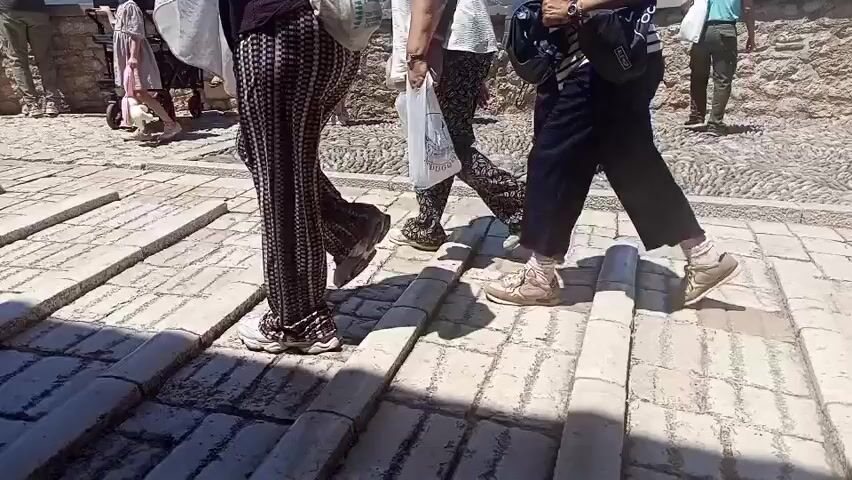 This video shows a group of people walking on a cobblestone path. The subjects are varied, including men, women, and what appears to be children. They are dressed in casual, summer clothing. One person in the foreground is wearing a patterned jumpsuit and the other in front is wearing black pants and a striped shirt.

The scene is outdoors, with a stone wall in the background. The ground is paved with cobblestones, and there are some raised stone elements that could be part of stairs or an architectural feature. The lighting suggests it is daytime, likely sunny given the shadows. The overall atmosphere appears to be relaxed and touristy, as people are out and about.

One person is carrying a white plastic bag with some writing on it, though it's not clearly legible. Another person is wearing a backpack. The people are moving in different directions, suggesting they are exploring the area or passing through. There are no overt displays of emotion, but the general impression is one of casual movement and sightseeing. The location, Mostar, Bosnia and Herzegovina, evokes images of historical architecture and popular tourist destinations.
