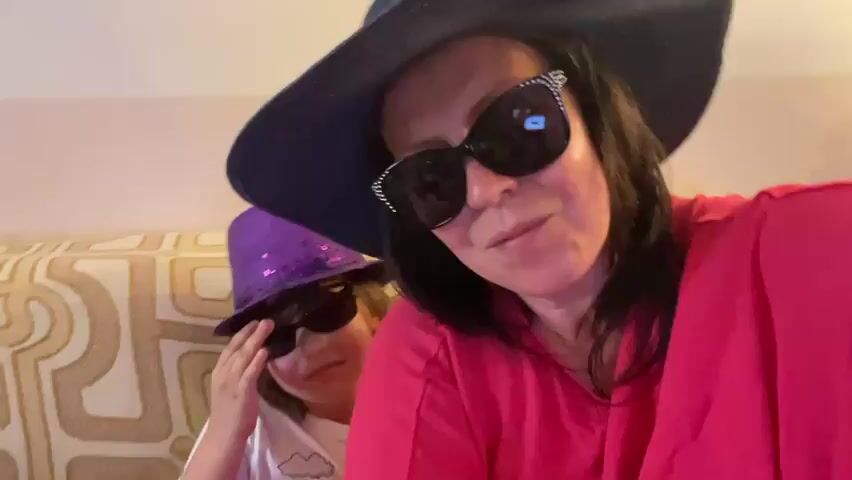 This is a short video clip, likely from a social media story or a personal recording. The primary subjects are two individuals: a woman and a child.

The woman is in the foreground, wearing a large, dark-brimmed hat and oversized sunglasses with decorative embellishments. She has dark hair and is wearing a bright pink top. She appears to be looking directly at the camera and smiling slightly, suggesting a playful or informal mood.

Behind her, to the left, is a child wearing a purple, sparkly top hat and dark sunglasses. The child's face is partially obscured, and their expression is difficult to discern clearly due to the lighting and the sunglasses. The child is reaching up towards their face with one hand.

They are both seated in what appears to be a living room or a similar indoor setting. Behind them, a patterned sofa or armchair is visible, with a cream-colored fabric featuring an abstract, geometric design in shades of beige and brown. The lighting is warm and artificial, suggesting it is likely daytime but indoors, or possibly evening.

The video exhibits some motion blur, particularly in the child's area and in brief moments across the entire frame, indicating movement and possibly a selfie-style recording where the camera operator is also moving. This movement adds to the candid and lively feel of the recording.

The overall impression is of a fun, lighthearted moment, possibly a dress-up or playful activity between the woman and the child. The prominent hats and sunglasses suggest a theme of disguise, performance, or simply embracing a whimsical mood. The setting and attire do not provide specific clues about the time of day or weather outside, but the indoor environment and the casual attire suggest a relaxed, at-home activity. No visible text is present in the media.