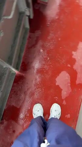 A person wearing blue pants and white shoes walks along a wet red deck of a boat. The deck is covered in puddles and water stains. A metal railing runs along the side of the deck, separating it from the water outside. In the distance, a wooded shoreline is visible under an overcast sky.
