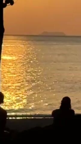 Two silhouetted figures sit on the sand at a beach, facing the ocean at sunset. The water reflects the golden light, creating a shimmering effect. An island is visible in the distance on the horizon. A tree is partially visible on the left side of the frame.