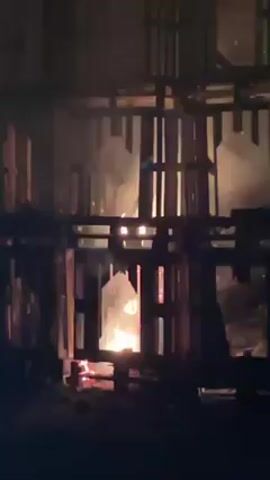 The video shows a structure made of wooden pallets engulfed in flames. The fire is concentrated at the base of the structure, with flames rising high inside the pallet enclosure. Smoke is visible, and there is ambient light suggesting it is nighttime. The wood is visibly burning, and the structure appears to be collapsing in places. There are some voices audible in the background.